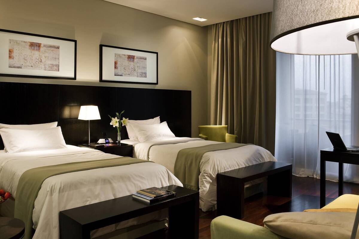 A hotel room with two beds, a desk with a laptop, a chair, a lamp, and a window with curtains.