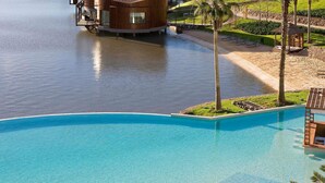 A large, clear blue pool with a wooden deck and lounge chairs, surrounded by a landscaped area with palm trees and a view of a body of water. - Sofitel La Reserva Cardales (Campana)