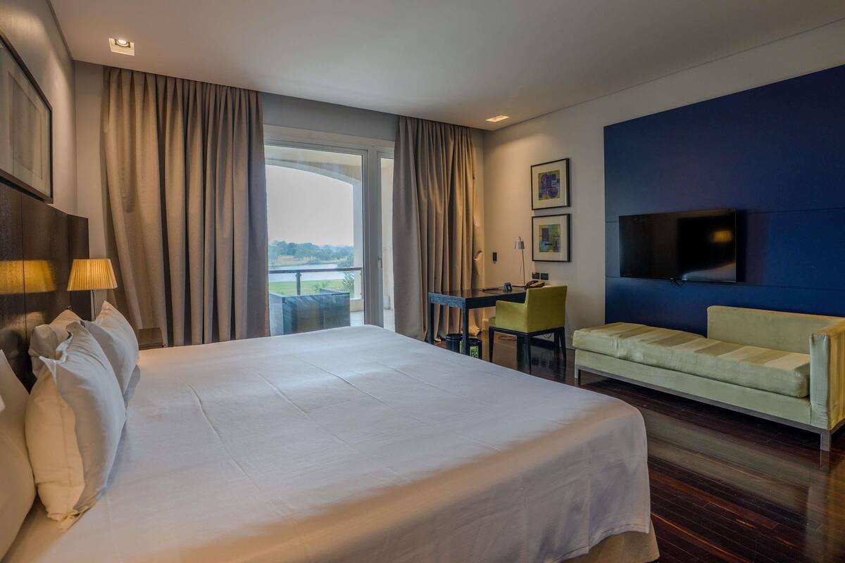 A hotel room with a large bed, a sofa, a desk with a chair, a TV mounted on the wall, and a view of the outdoors through a large window.