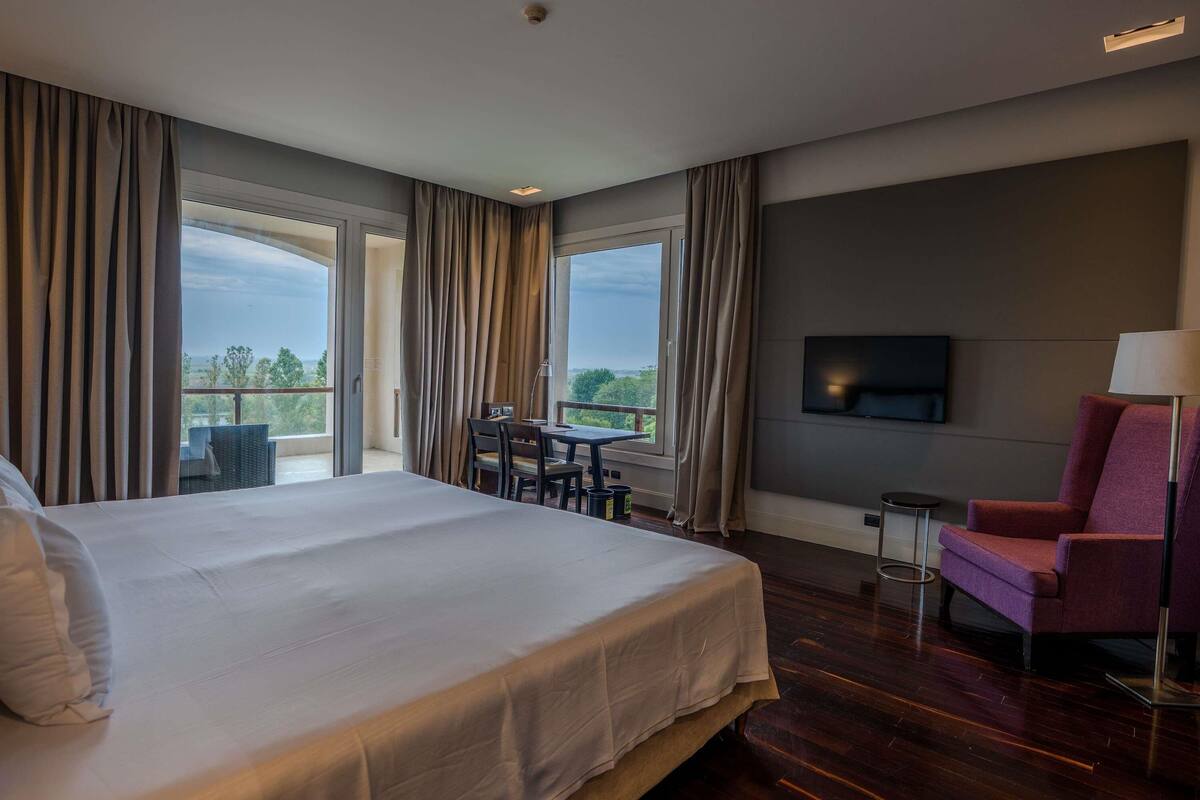 A hotel room with a large bed, a desk with chairs, a TV mounted on the wall, and a balcony with curtains.
