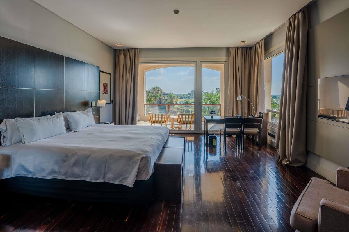 A hotel room with a large bed, a desk, a chair, and a balcony with a view.