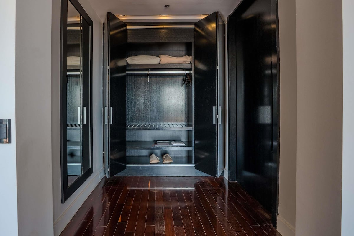 A hallway with a built-in wardrobe featuring a mirrored closet door, a shelf with shoes, and a bed with a headboard.