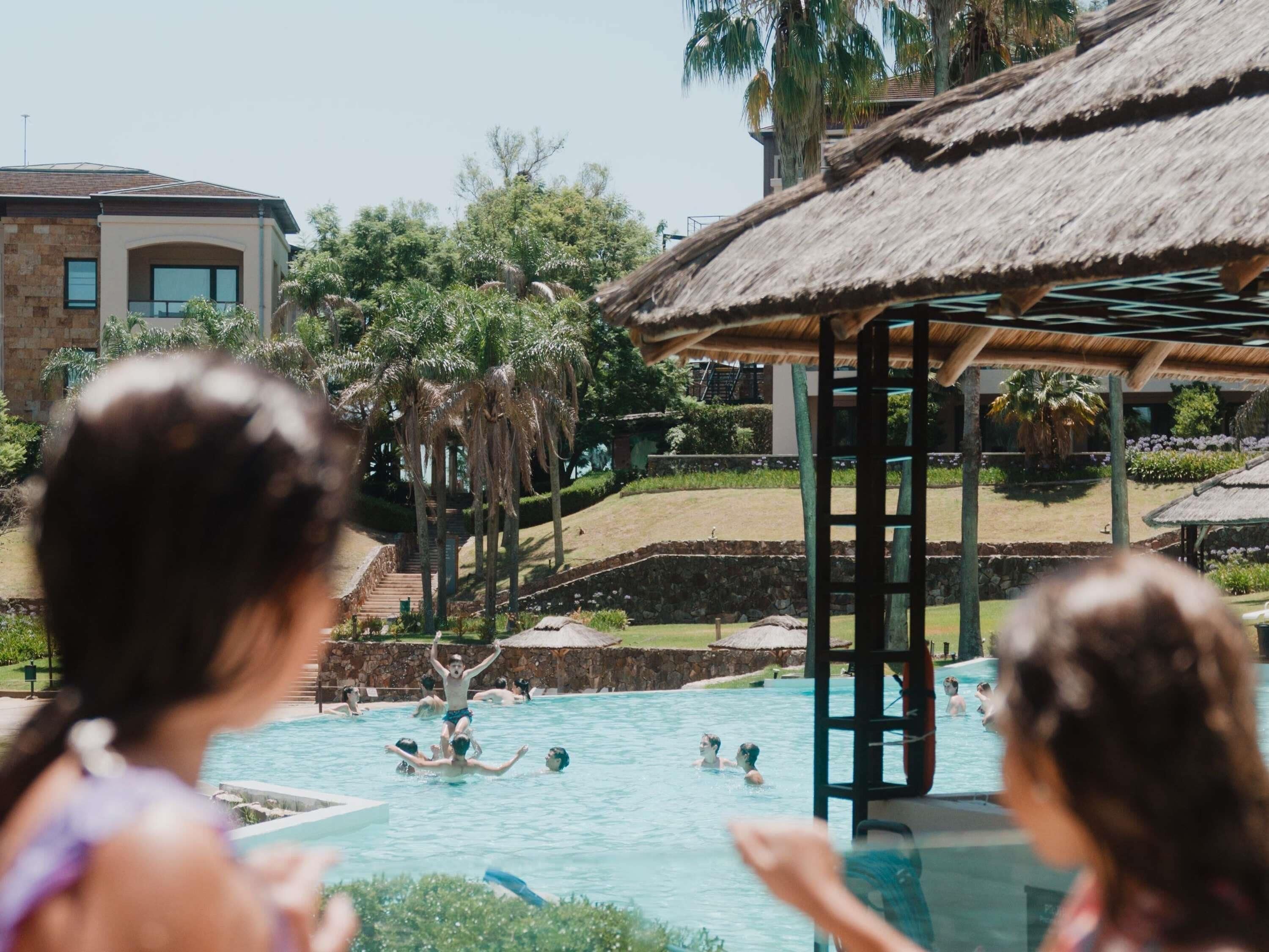 A swimming pool area with a thatched-roof structure, palm trees, and a stone wall.
