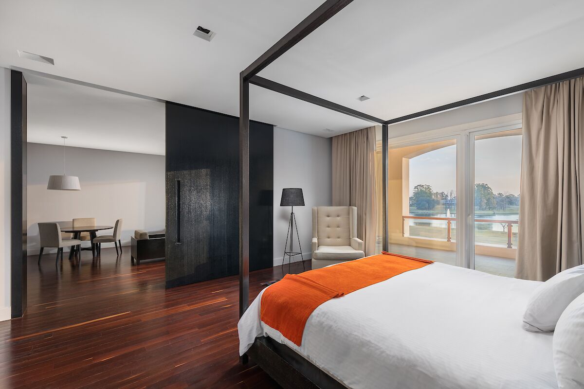 A modern hotel room with a large bed, a dining area, and a view of the outdoors.