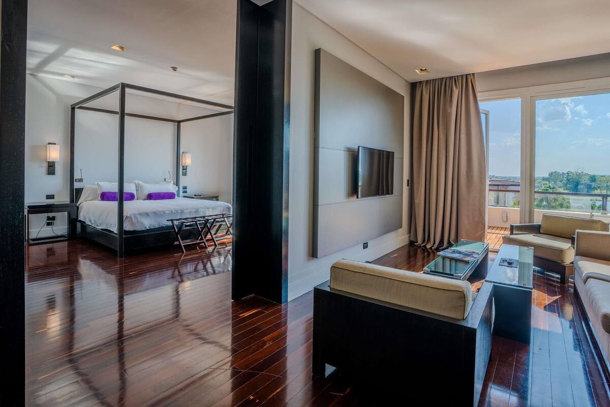 A modern hotel room with a four-poster bed, a flat-screen TV mounted on the wall, a balcony with a view, and a glass coffee table.