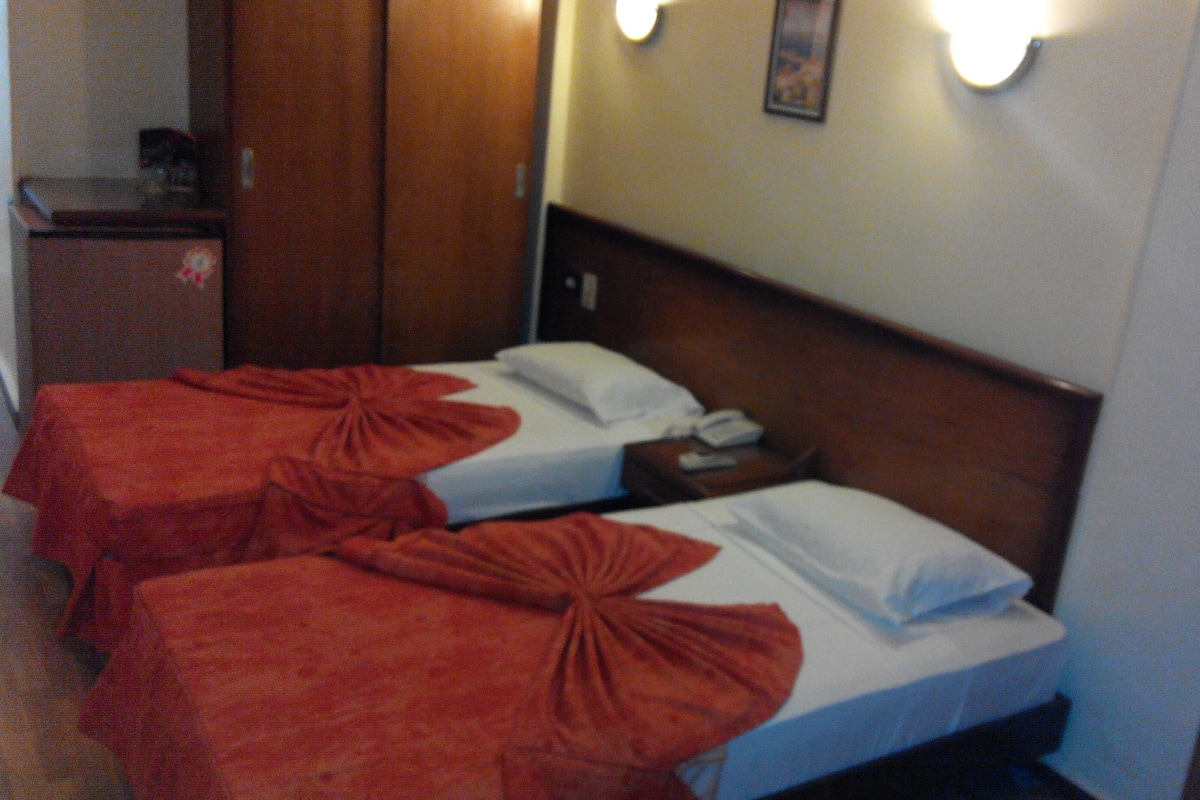 A hotel room with two beds, each with a red blanket and white pillow. There is a nightstand with a phone and a lamp on it.