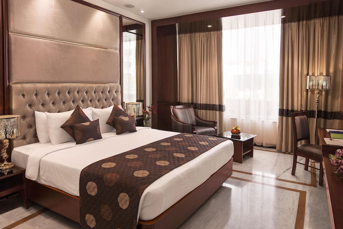 A spacious hotel room with a large bed, a sitting area with a chair, a desk, and a large window with curtains.