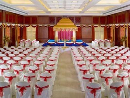 Un salón de conferencias con filas de sillas blancas adornadas con cintas rojas, un escenario con un telón de fondo azul y rojo, y un dosel dorado.