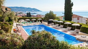 Piscina con sillas de descanso y sombrillas, rodeada de vegetación y con vista a un pueblo costero.