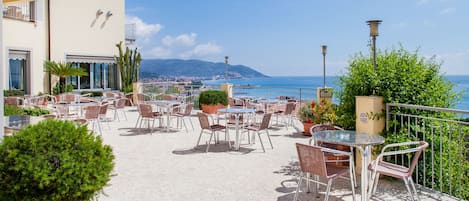 An outdoor café with tables and chairs, potted plants, and a view of the sea.