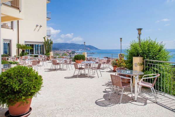 An outdoor café with tables and chairs, potted plants, and a view of the sea. - Hotel Metropol (Diano Marina)