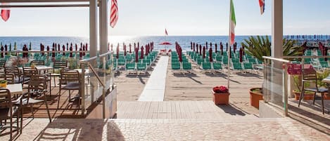 An outdoor seating area with a view of the sea, featuring a tiled floor, chairs, tables, and potted plants.