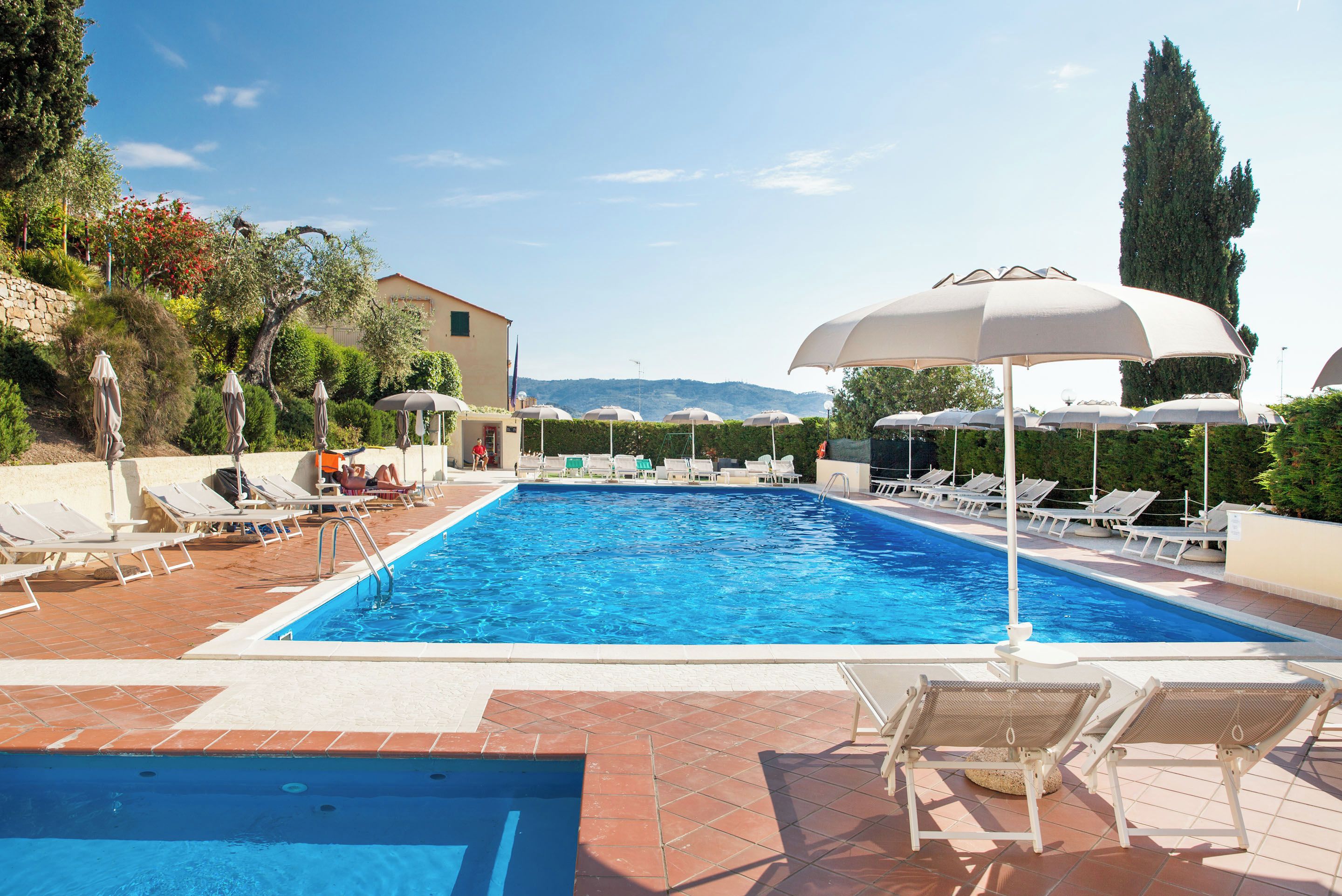 A swimming pool with sun loungers and umbrellas, surrounded by a landscaped garden and a building.