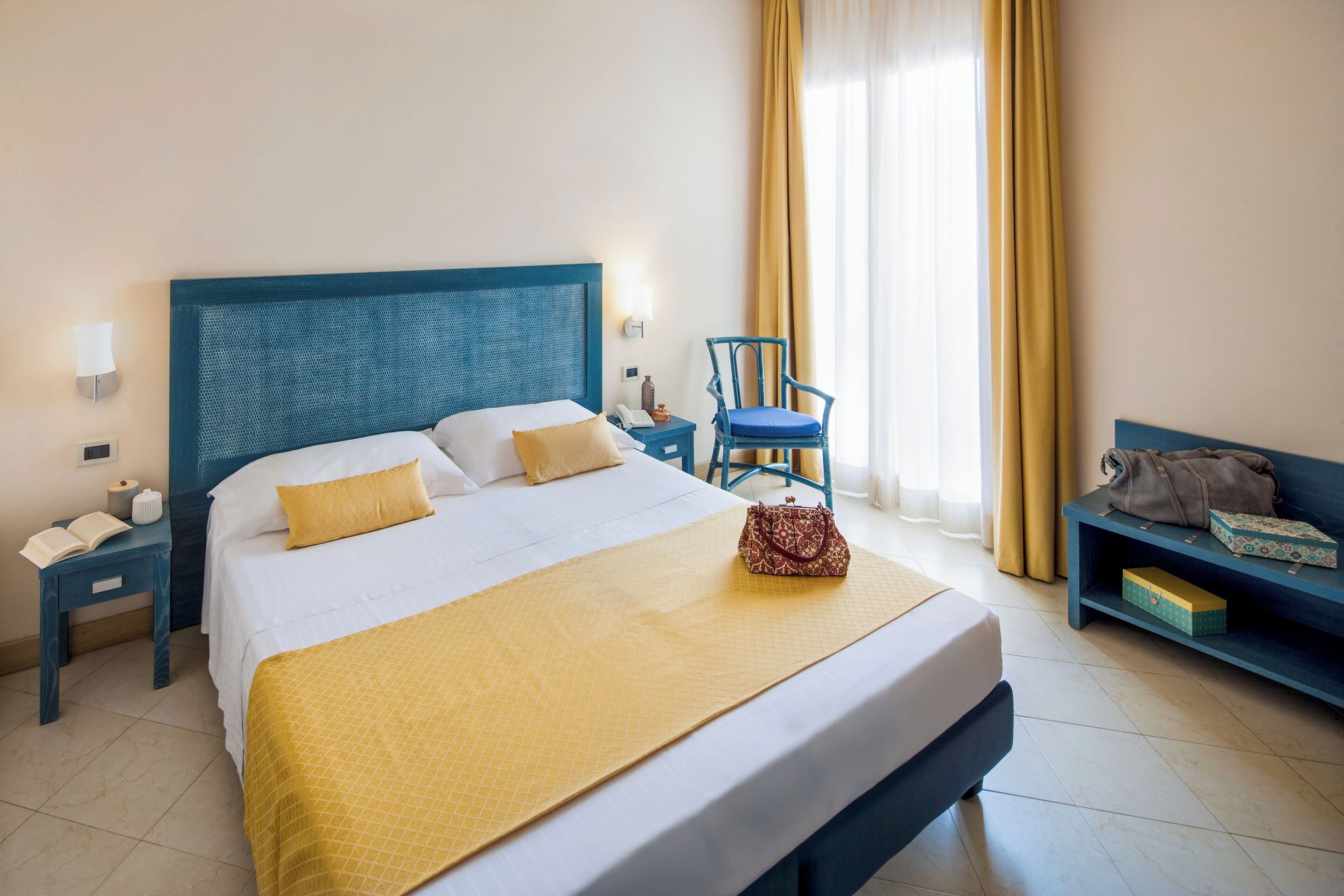 Quarto de hotel com cama grande, mesa de cabeceira com abajur, cadeira e um banco com uma mala e caixa.