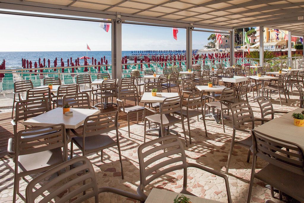 En udendørs café med borde og stole, med udsigt over en strand hvor folk er i det fjerne.