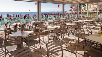 Un café al aire libre con mesas y sillas, con vista a una playa donde se ven personas a lo lejos.