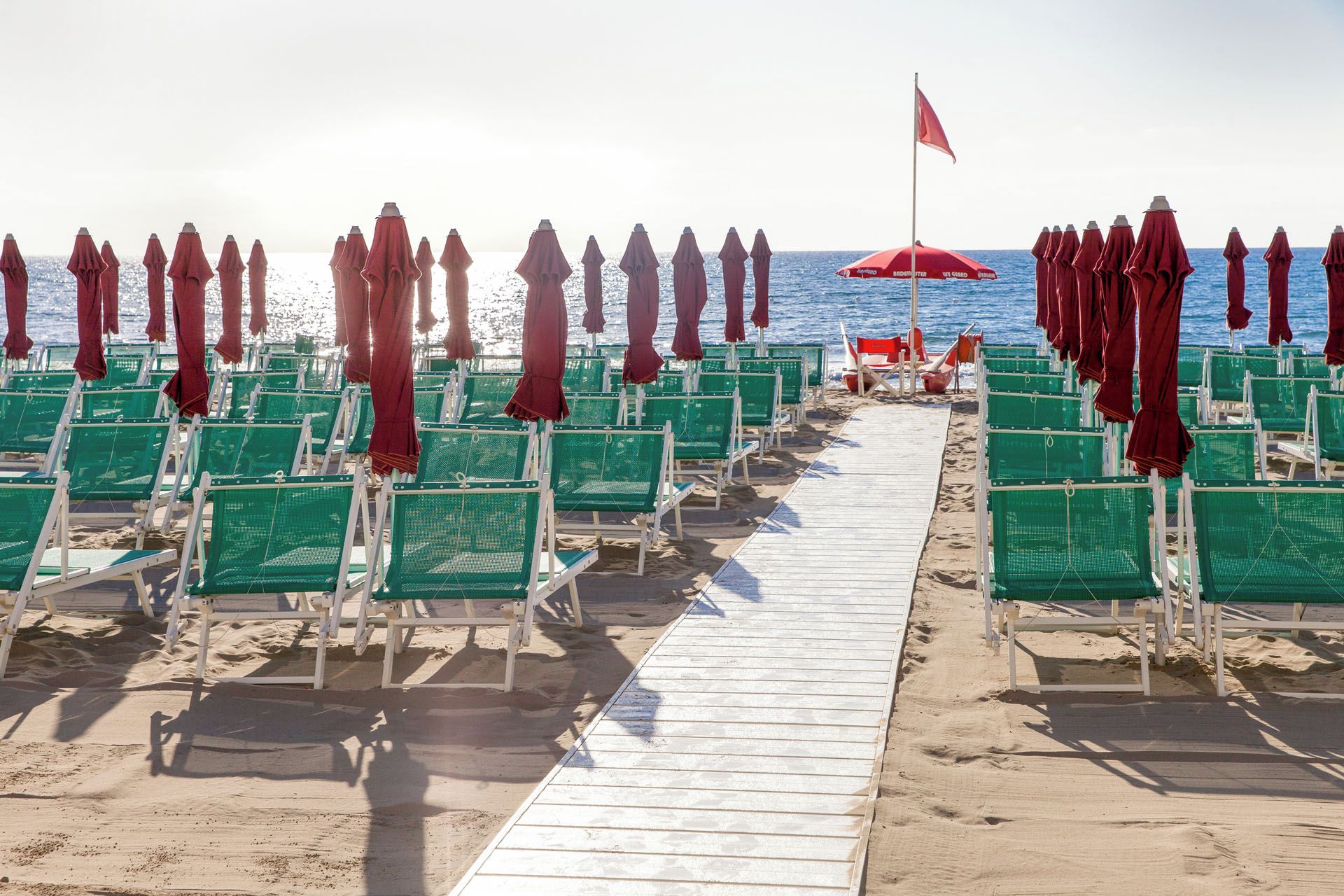 Una playa con filas de sillas verdes y sombrillas rojas, un paseo de madera y una bandera.