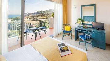 Quarto de hotel com varanda, cama, escrivaninha com cadeira, televisão e vista para a cidade e o mar.