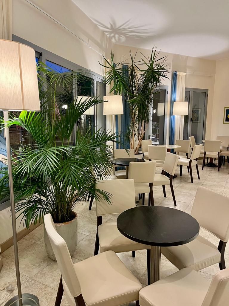 En hotellobby med runda bord och stolar, stora krukväxter och en modern inredningsstil.