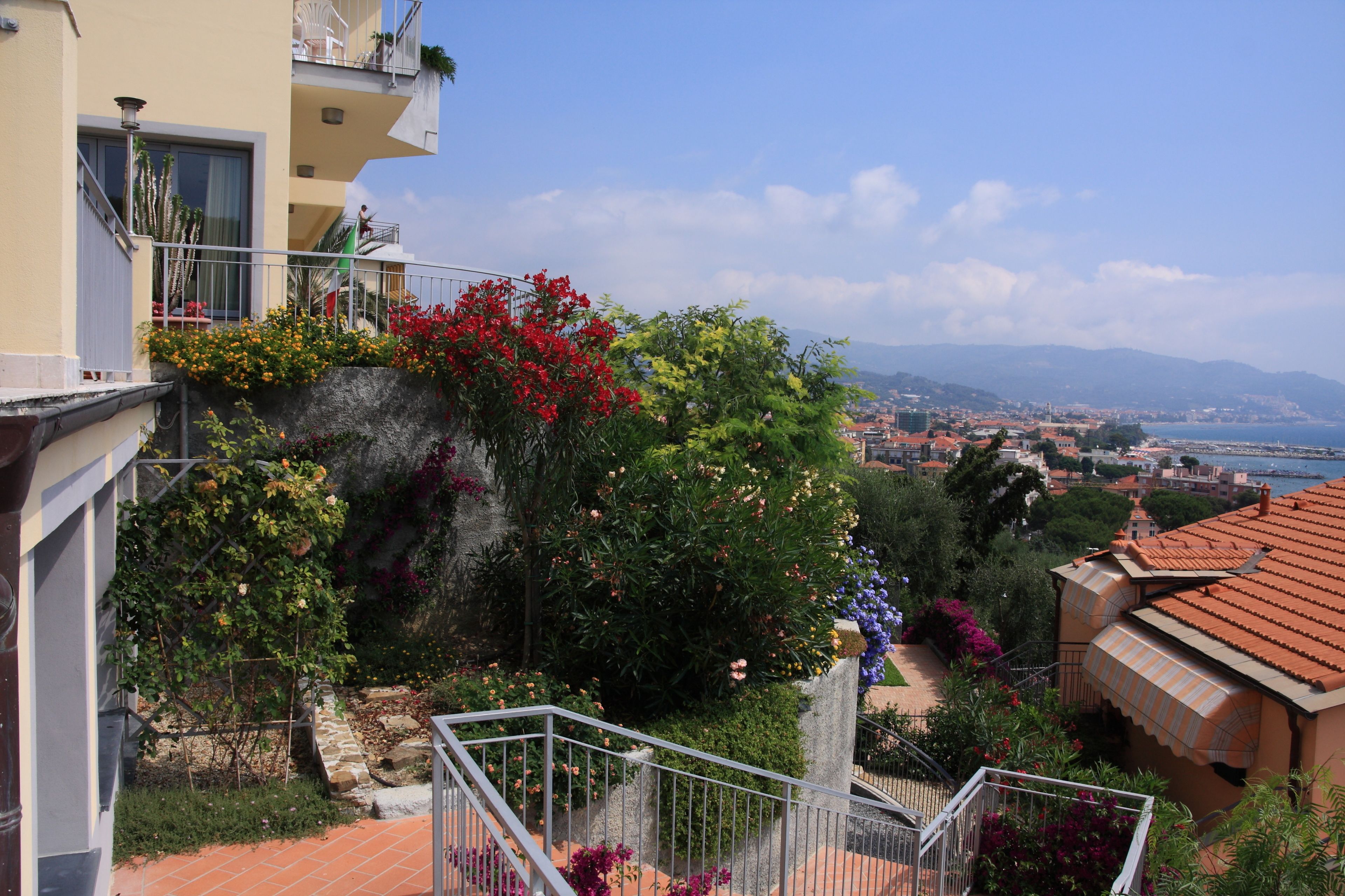 A residential building with a balcony, a garden with various plants and flowers, and a view of a coastal town.