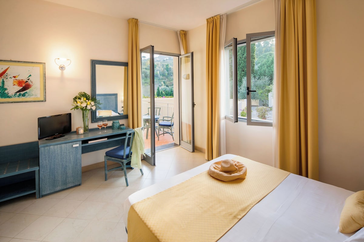 Habitación de hotel con cama, escritorio con computadora, una silla y balcón con vistas a la vegetación.