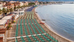 Una playa con hileras de sombrillas verdes y tumbonas, una orilla arenosa y edificios al fondo.