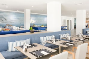 A modern dining area with blue seating, tables set with cutlery, and a large window with a view. - Reverence Mare Hotel - Adults Only (Calvia)