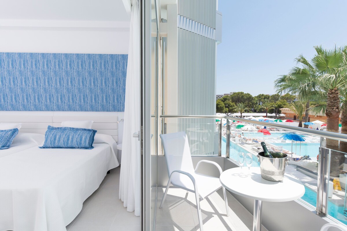 A balcony with a pool view, a bed with white bedding, and a table with a champagne bottle.