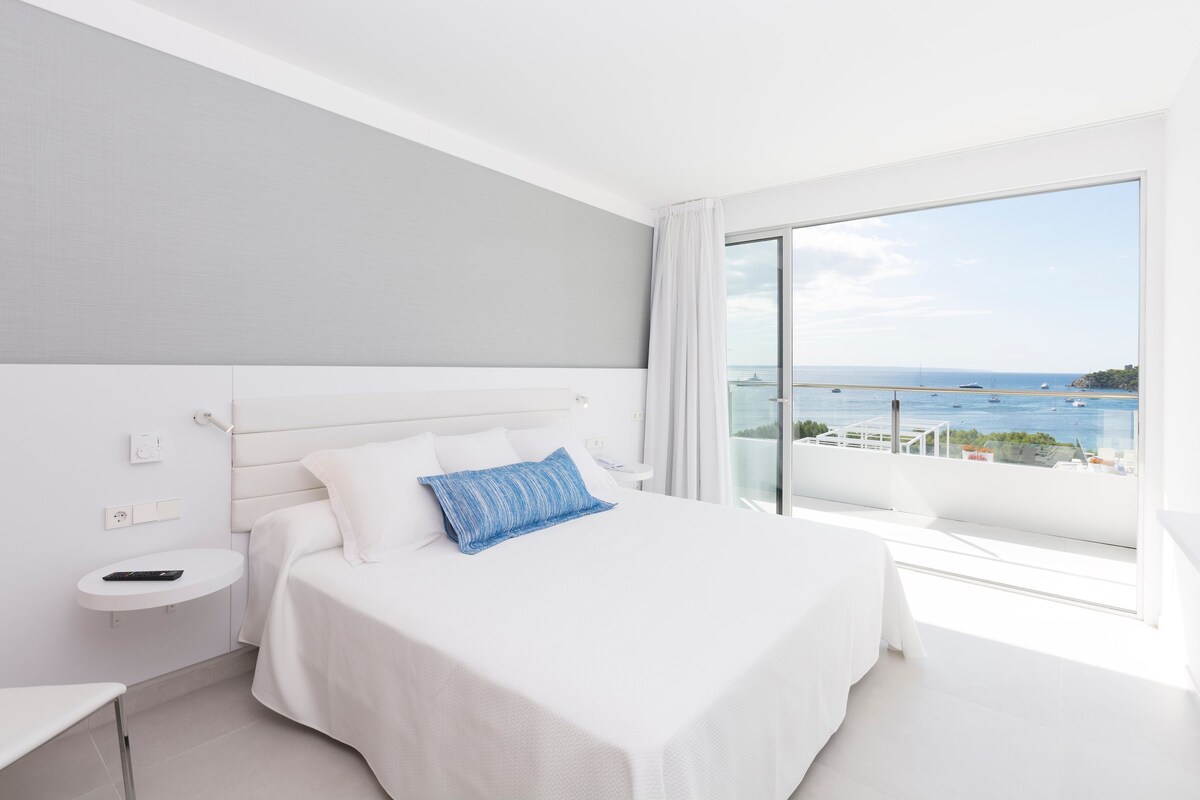 A hotel room with a large bed, a bedside table, a remote control, and a view of the sea.