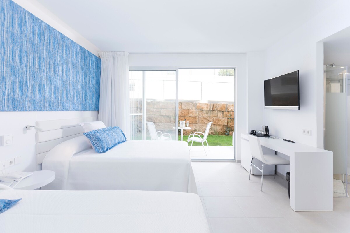 A modern hotel room with two beds, a desk, a TV, and a balcony with outdoor seating.