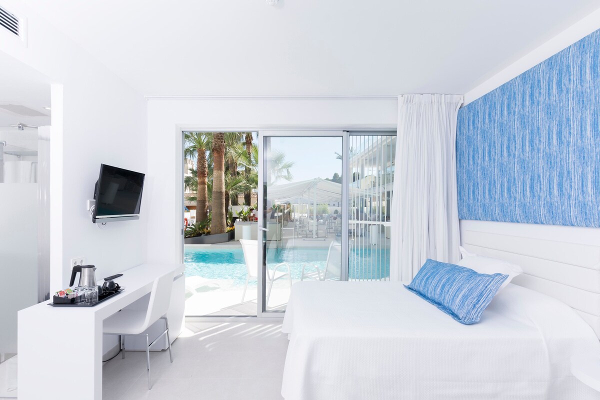 A modern hotel room with a bed, a desk with a flat-screen TV, a pool view, and a sliding glass door.