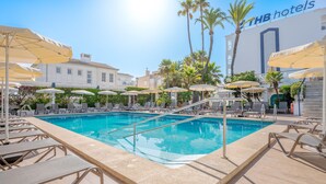 Un espace piscine avec des chaises longues et des parasols, une piscine à l’eau d’un bleu limpide et, au fond, le bâtiment de l’hôtel.