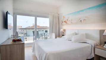 Una habitación de hotel con una cama grande, un escritorio con fruta, una silla, un balcón con vistas a edificios y un mural temático de playa.