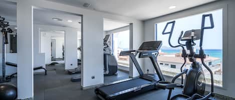 Une salle de sport moderne équipée de tapis de course et de vélos elliptiques, de grandes fenêtres offrant une vue sur la mer et d’un aménagement spacieux.