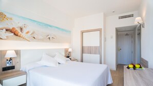 Une chambre d’hôtel avec un lit, une table de chevet, une lampe, un miroir fixé au mur et un panier de fruits posé sur une table.