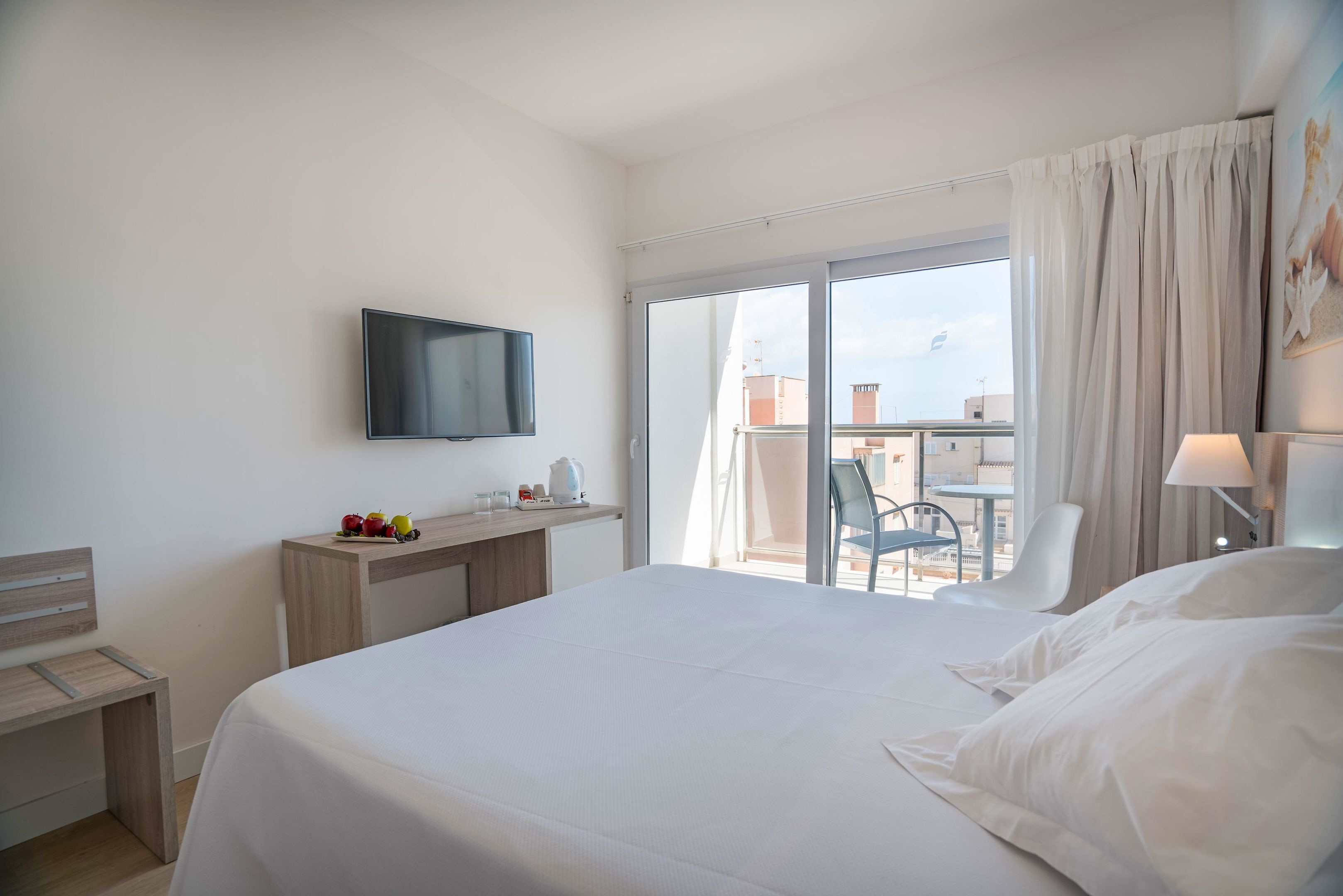 Een hotelkamer met een bed, een bureau met een flatscreen televisie, een balkon met stoelen en uitzicht op de stad.