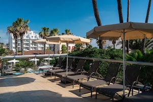 Zona de piscina con sillones y sombrillas, vista despejada de la alberca y un edificio con balcones al fondo.