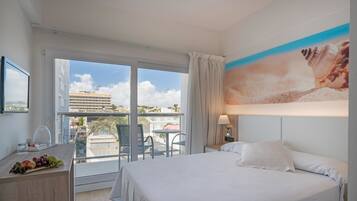 Ein Hotelzimmer mit einem Bett, einem Schreibtisch mit Obstkorb, einem Sessel und Blick auf die Stadt durch große Fenster.