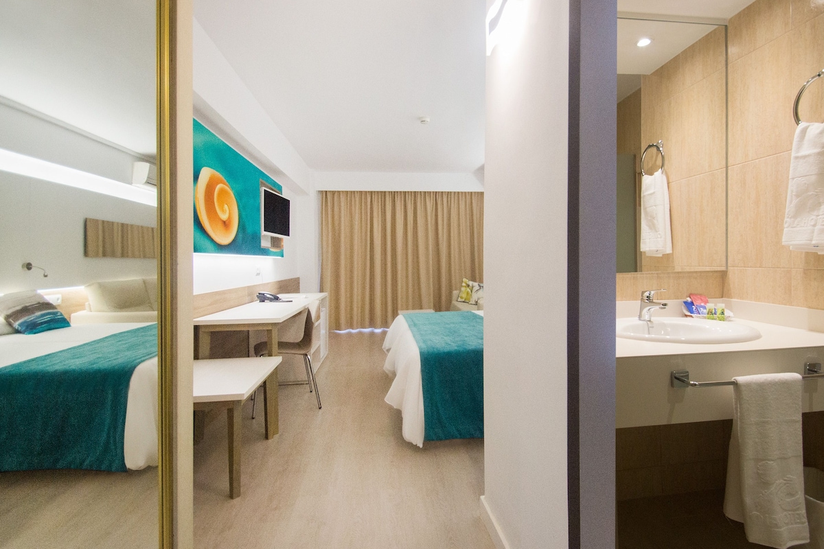 A hotel room with two beds, a desk, a chair, a TV, a bathroom with a sink and mirror, and a large mirror.