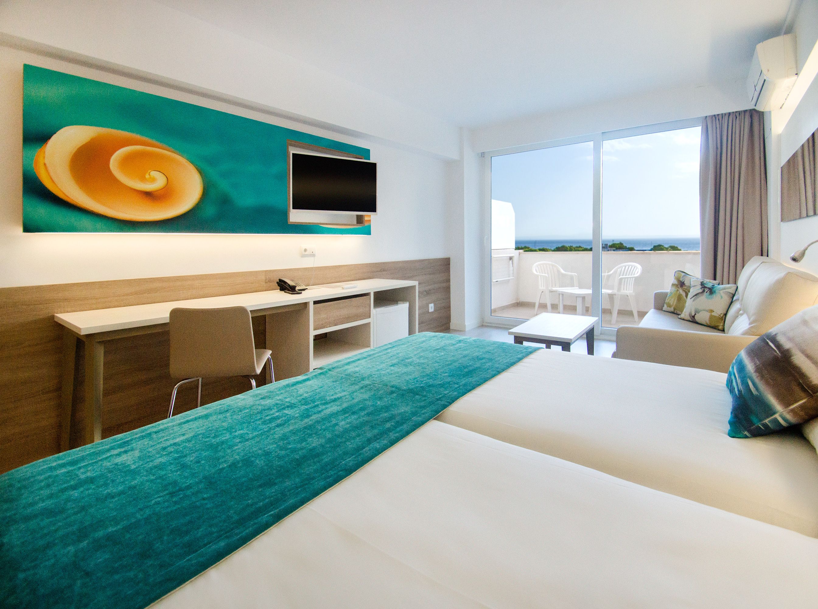 Habitación de hotel con cama, escritorio, silla, televisor y balcón con vista al mar.