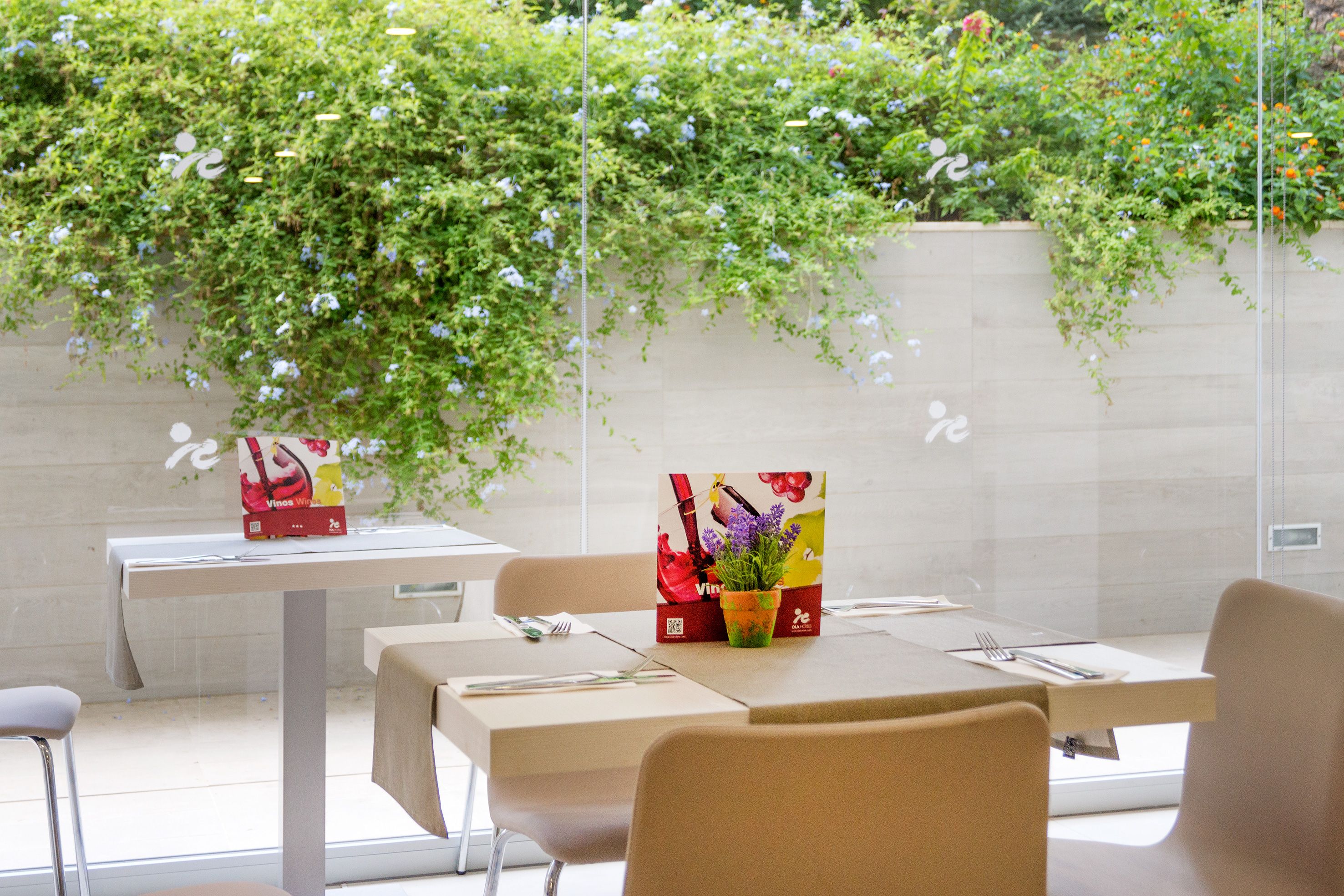 A restaurant with tables set for dining, a potted plant on the table, and a wall with climbing plants.