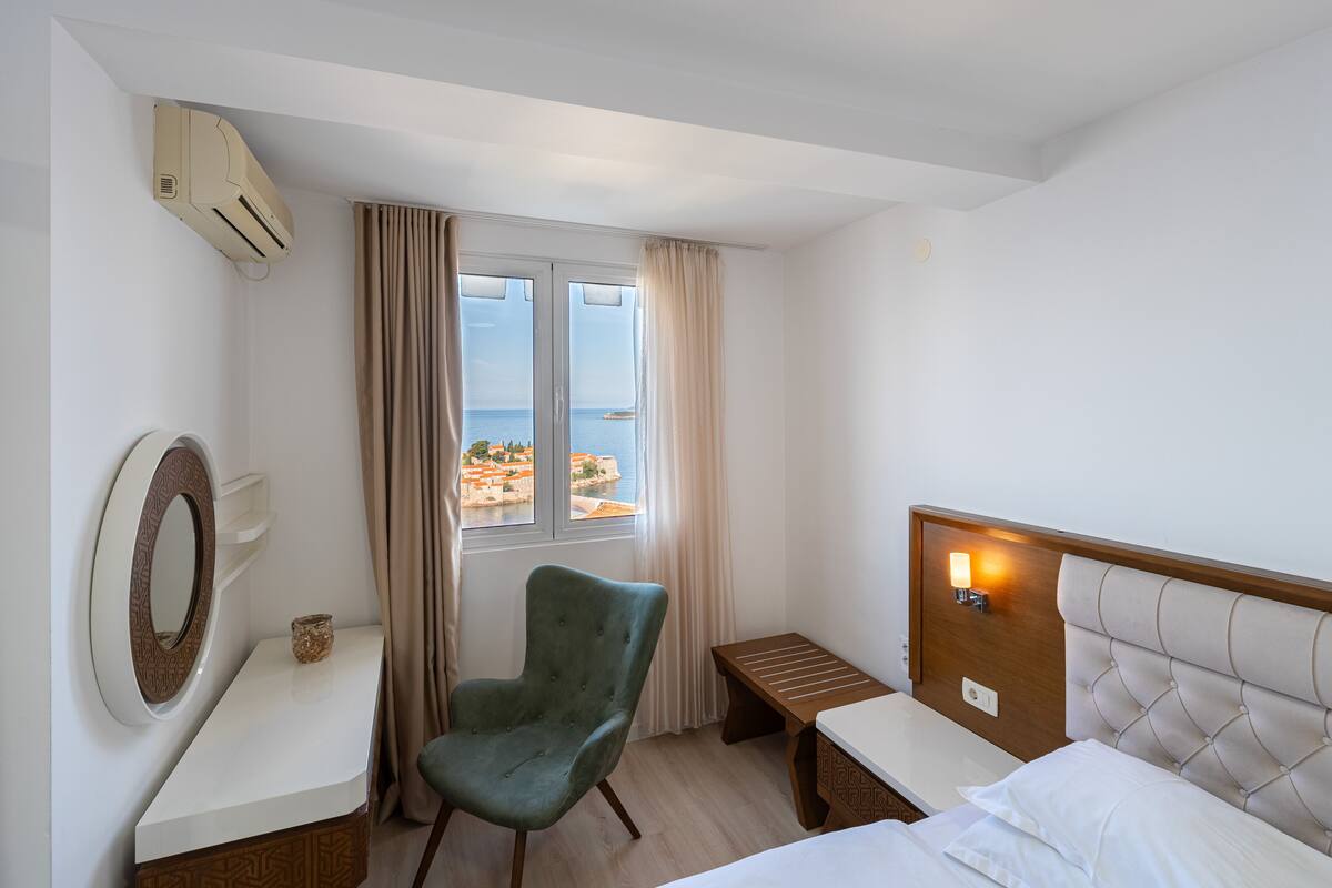 Une chambre d’hôtel avec un lit, une chaise, une table de chevet, un miroir et une fenêtre avec des rideaux.