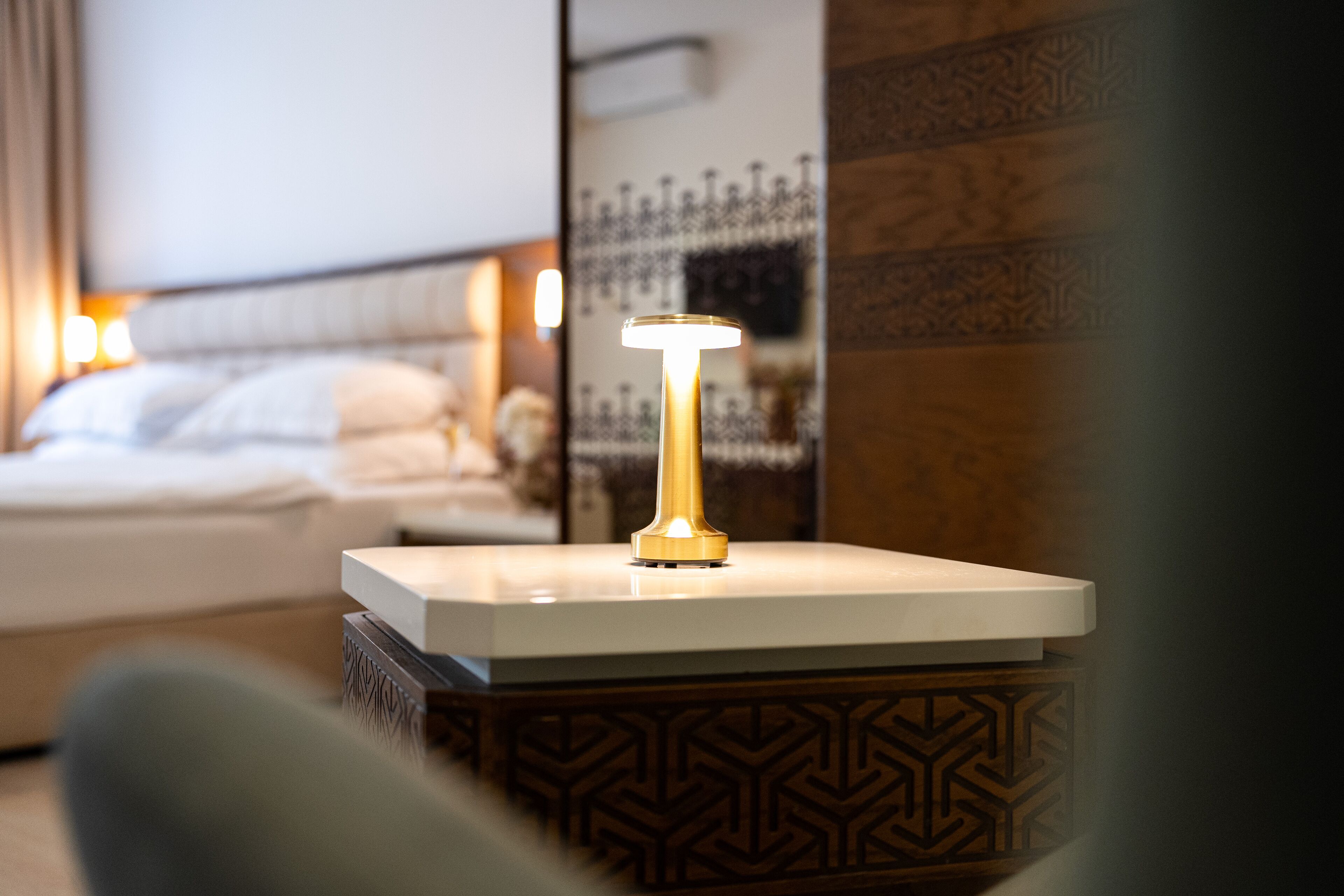 Un comodino con una lampada moderna, un angolo notte decorato e un letto con lenzuola bianche in una camera d'hotel.
