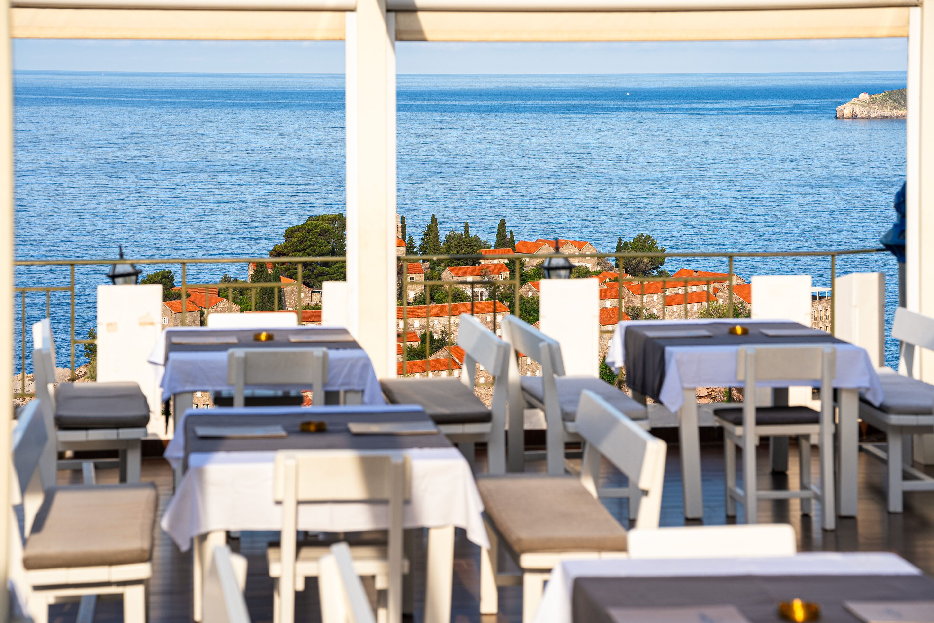 An outdoor dining area with tables and chairs, overlooking a coastal town and the sea.