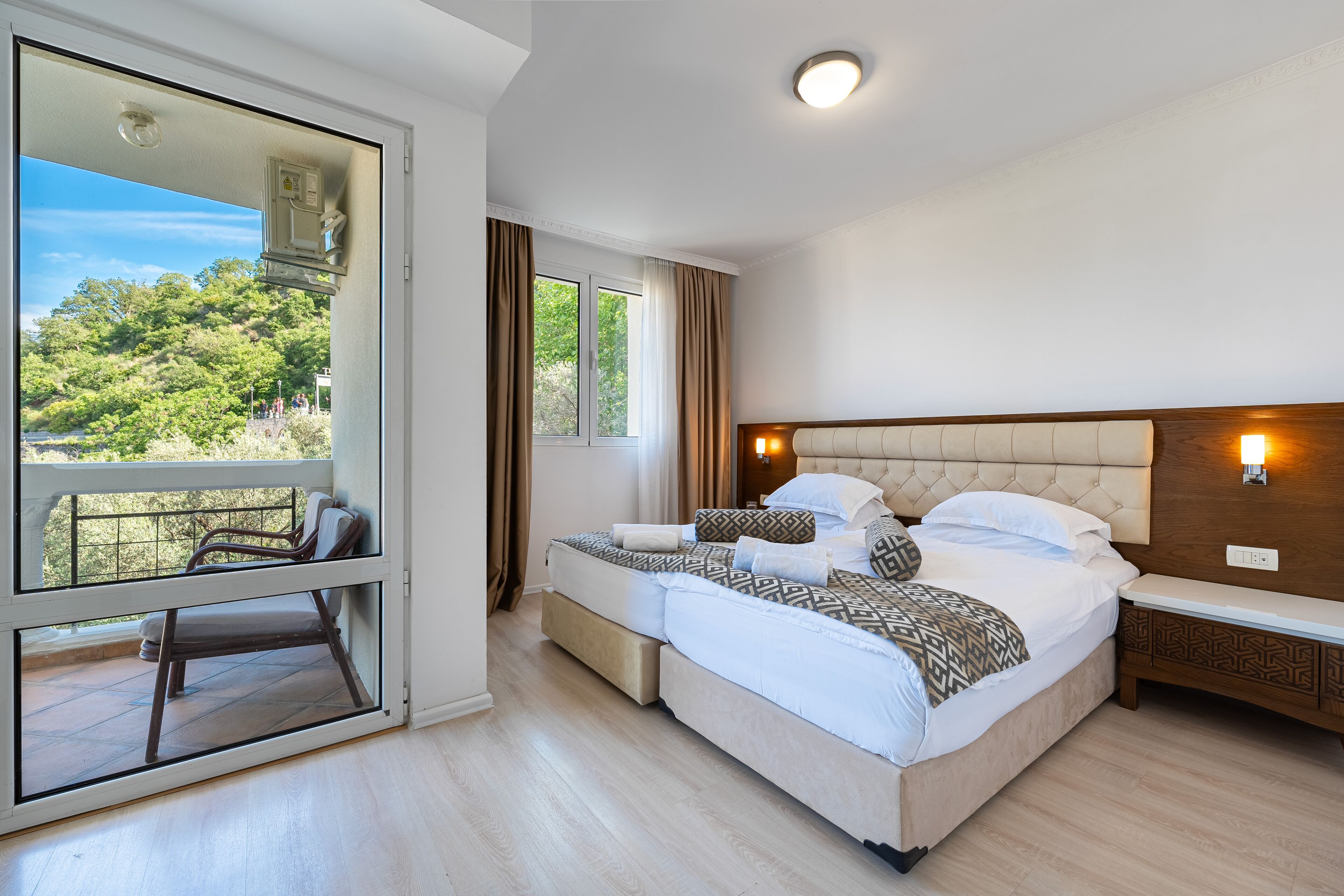Un dormitorio con una cama grande, una mesita de noche, un espejo y vistas al verdor exterior.