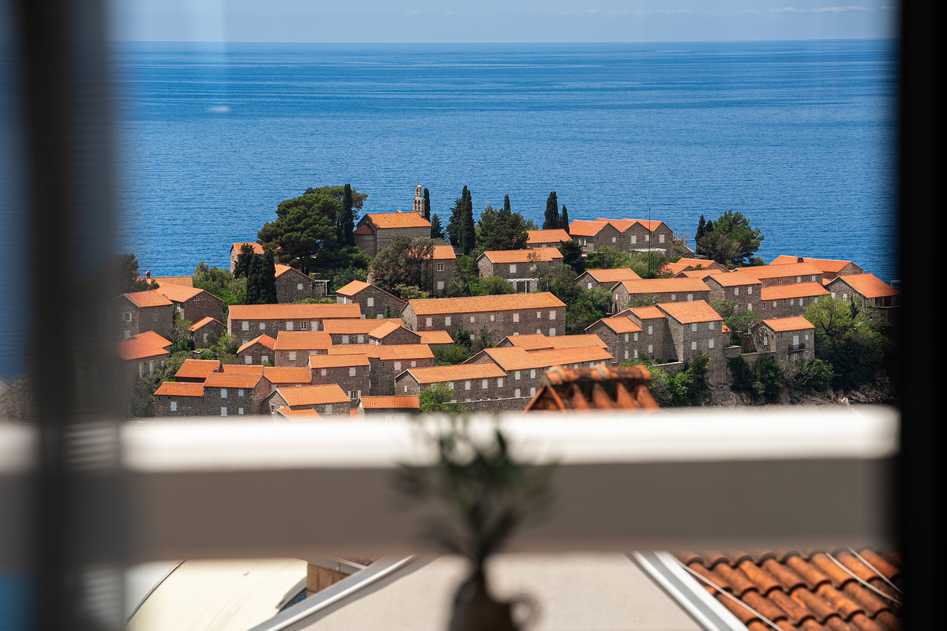 Ein Küstendorf mit terrakottafarbenen Dächern, freiem Blick auf das Meer und einem kleinen Baum im Vordergrund.
