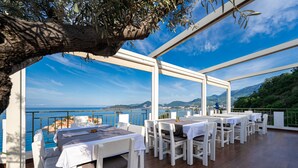 Un área de comedor al aire libre con mesas y sillas blancas, con vista al mar y las colinas.