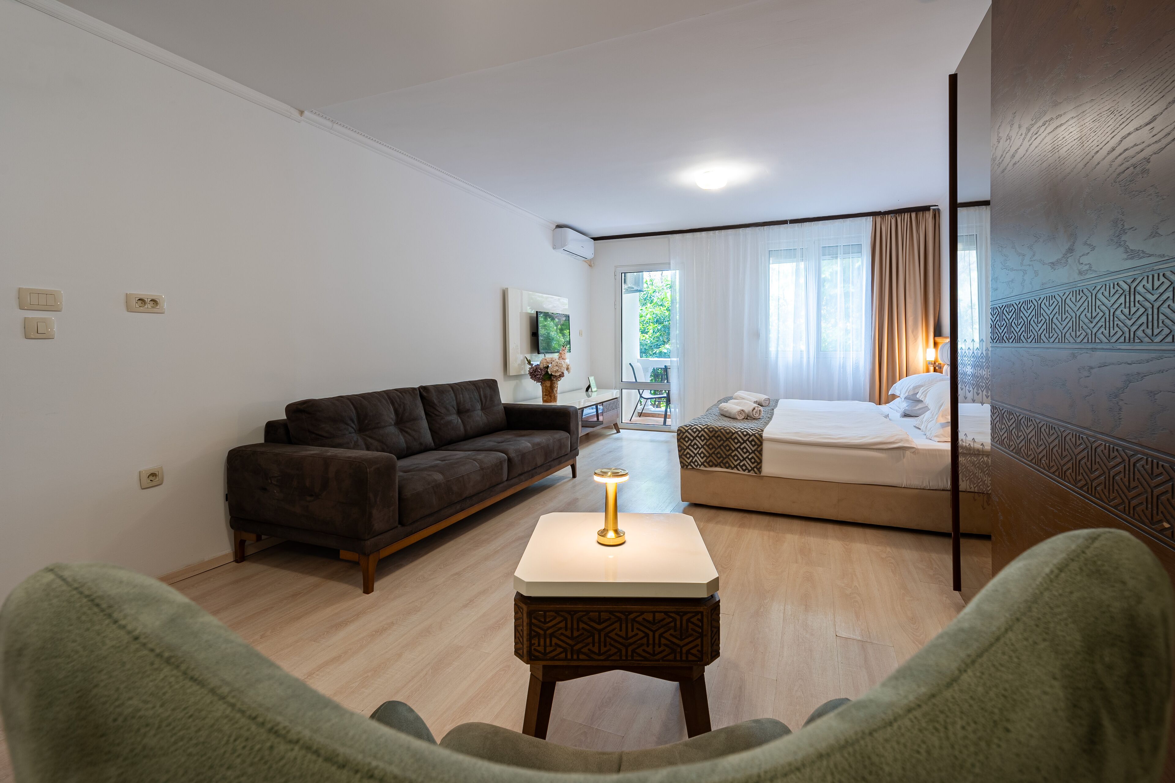 Moderni hotellihuone, jossa on sohva, pieni pöytä lampun kanssa, verhoilla varustettu sänky ja näkymä ulos.