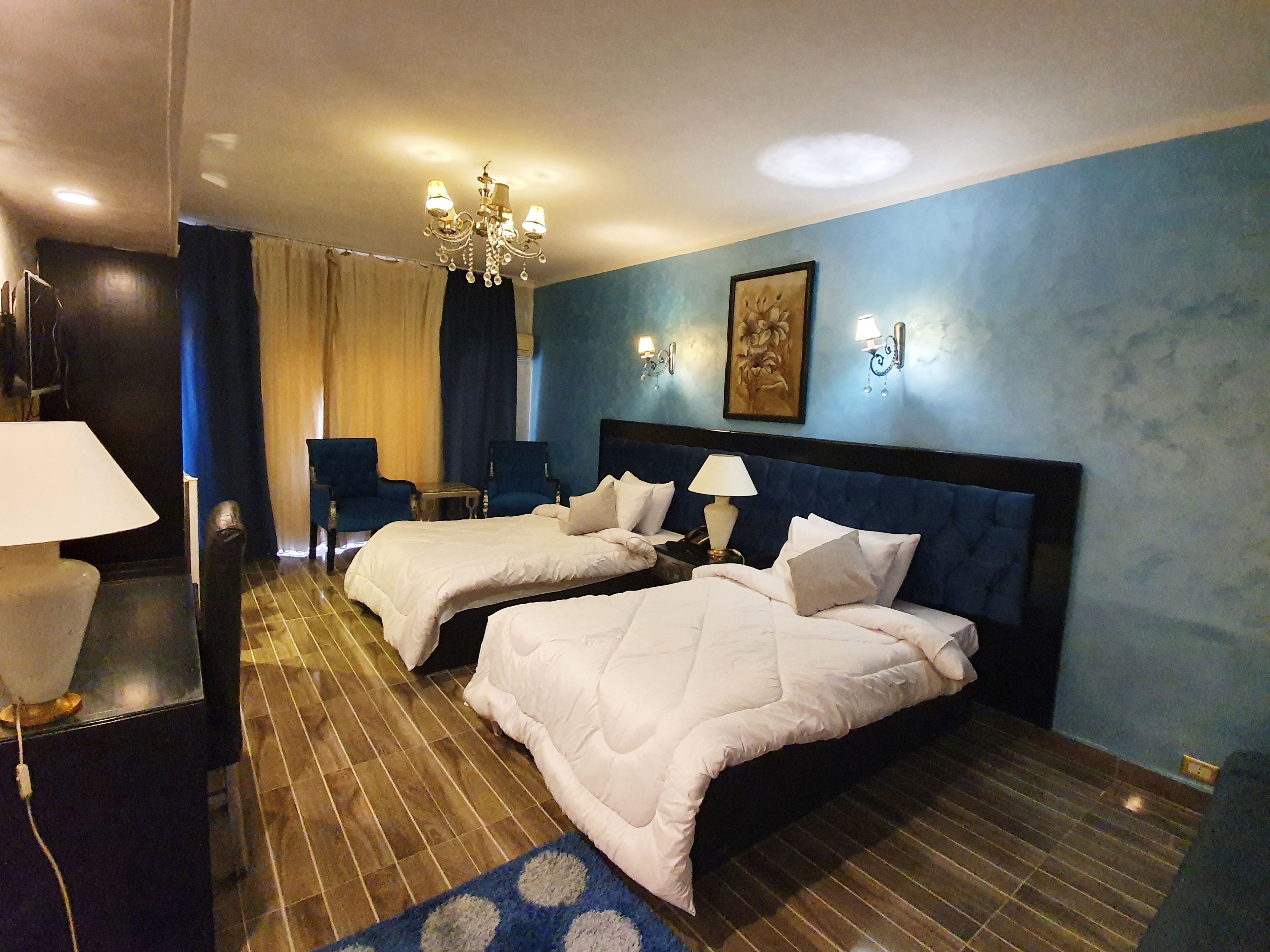 A hotel room with two beds, a chandelier, a desk with a lamp, and a blue wall with a painting.