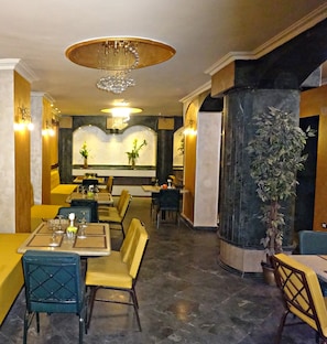 Un interior de restaurante con mesas, sillas, una araña y una columna grande con una planta.